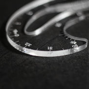 A close-up of a clear plastic protractor with angle markings, resting on a dark textured surface.
