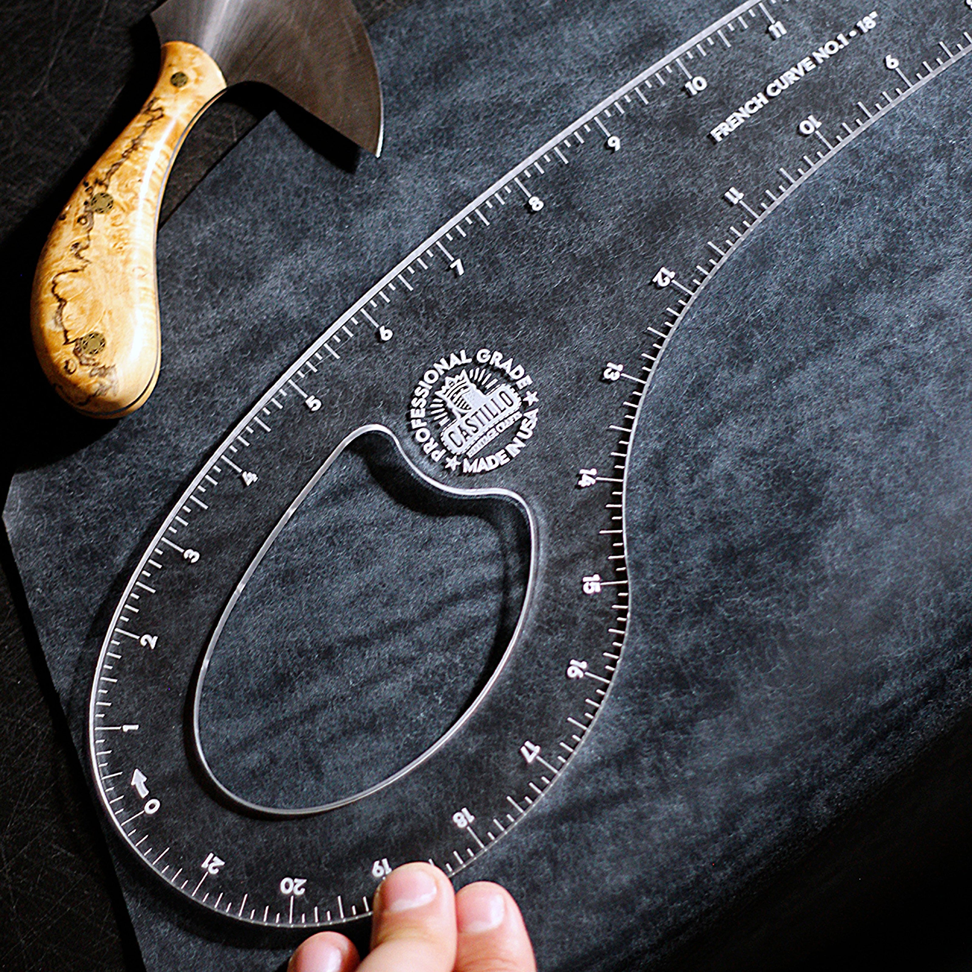 A clear French curve ruler with engraved measurement markings and a circular center hole, shown on a dark textured surface. A hand is holding the lower edge, and part of a wooden-handled tool is visible in the upper left.