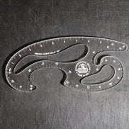 A clear acrylic drafting ruler with multiple curved sections, featuring black printed measurement markings and a circular Castle brand logo, photographed on a dark textured metal surface.