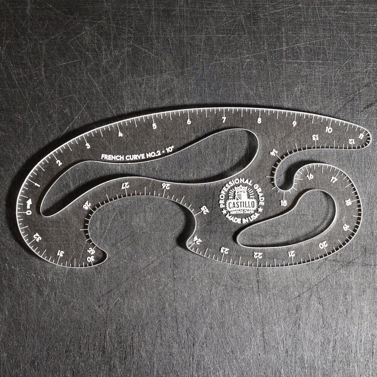 A clear acrylic drafting ruler with multiple curved sections, featuring black printed measurement markings and a circular Castle brand logo, photographed on a dark textured metal surface.