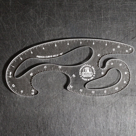 A clear acrylic drafting ruler with multiple curved sections, featuring black printed measurement markings and a circular Castle brand logo, photographed on a dark textured metal surface.