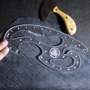 A clear plastic French Curve No. 3 by Castiron shown on a dark textured surface, with a hand holding one end and a carving knife with a light wood handle placed above it.