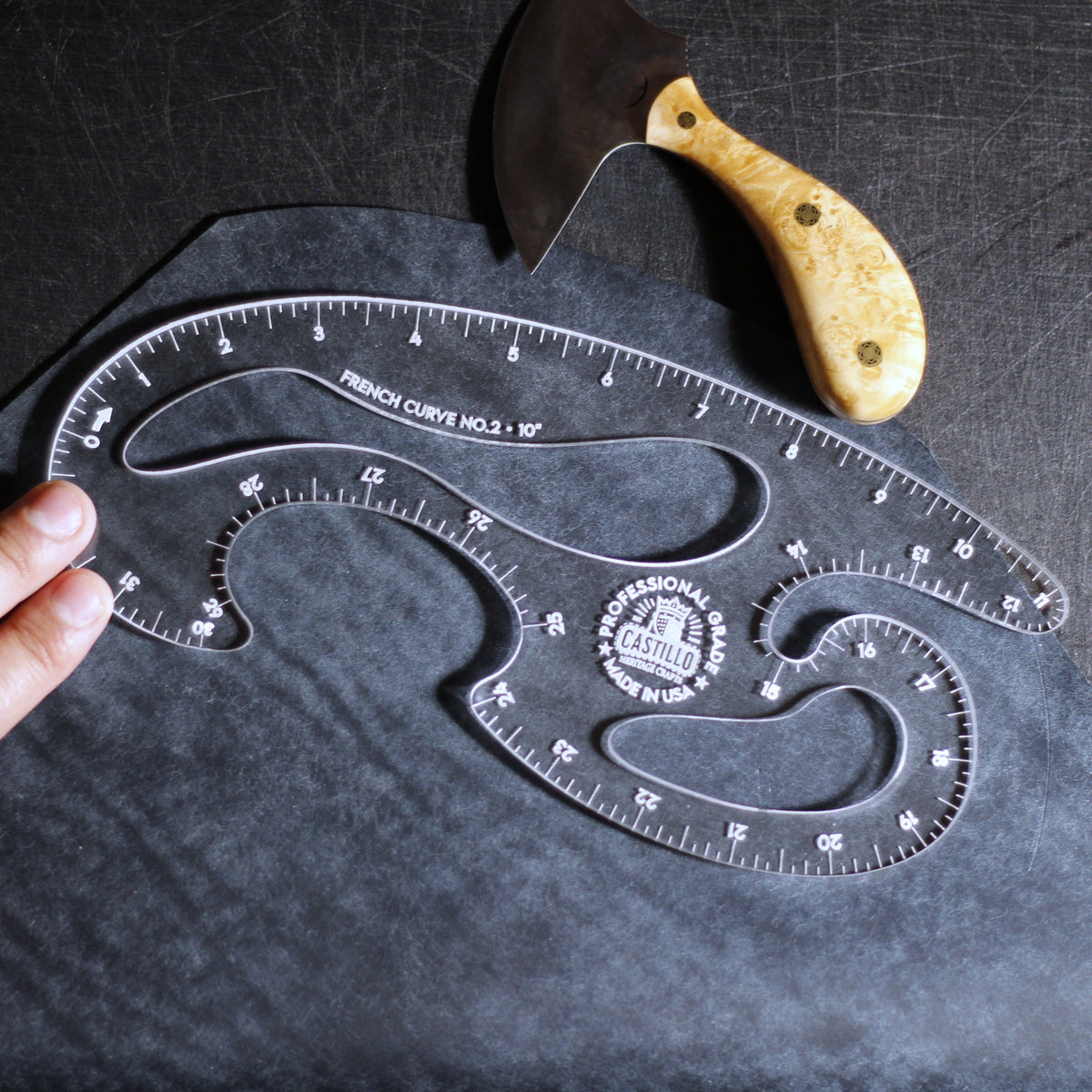 A clear plastic French Curve No. 3 by Castiron shown on a dark textured surface, with a hand holding one end and a carving knife with a light wood handle placed above it.