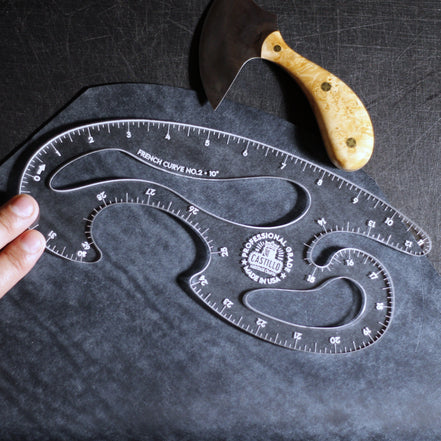 A clear plastic French Curve No. 3 by Castiron shown on a dark textured surface, with a hand holding one end and a carving knife with a light wood handle placed above it.