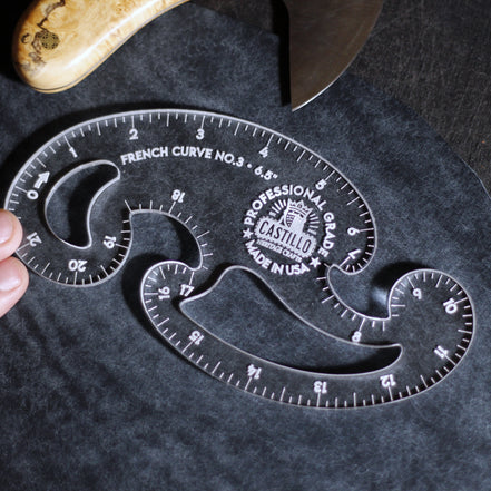 A clear acrylic French curve ruler, model No. 3 by Castillo, shown being held by a hand on a dark textured surface with a knife partially visible in the background.
