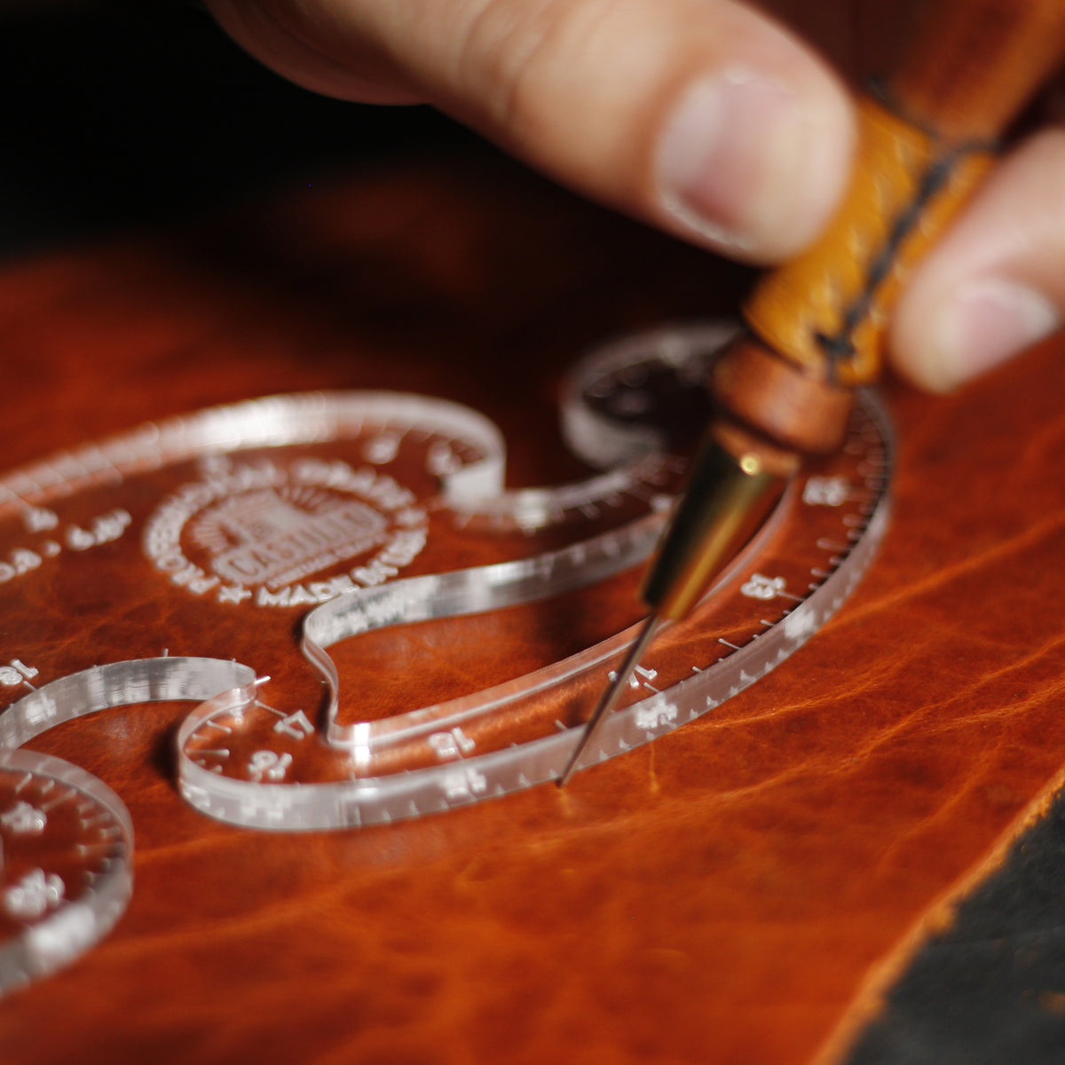 A close-up of a leatherworking scene showing a hand using a clear acrylic template with engraved markings over a rich brown leather surface, while holding a wooden-handled tool.