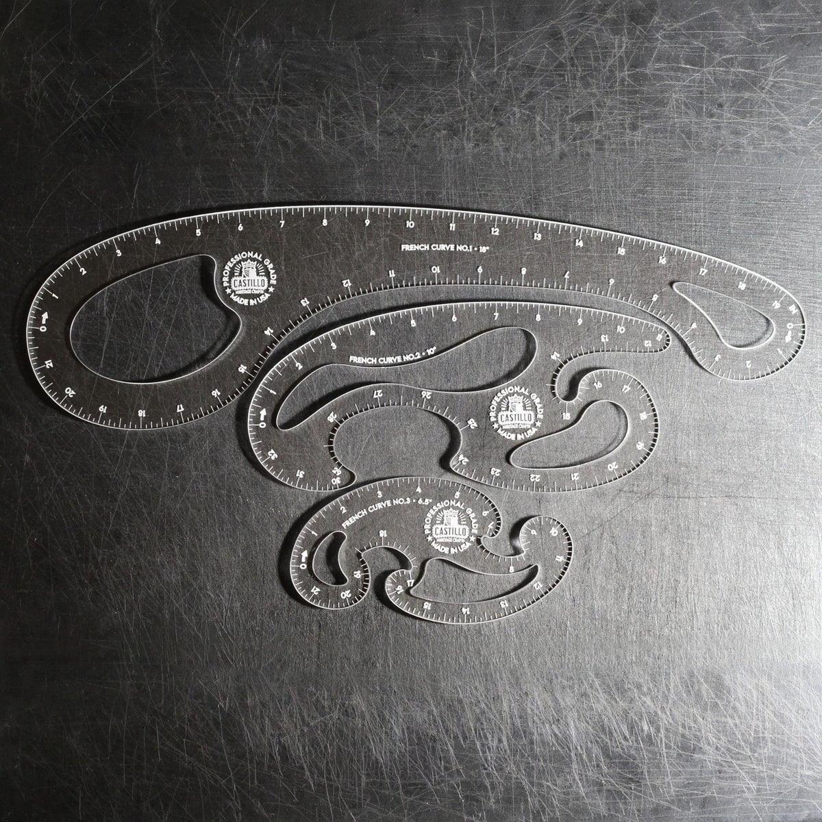 Three clear plastic French Curve Ruler sets of different sizes arranged on a dark, scratched metal surface.