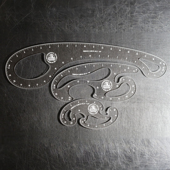 Three clear plastic French Curve Ruler sets of different sizes arranged on a dark, scratched metal surface.